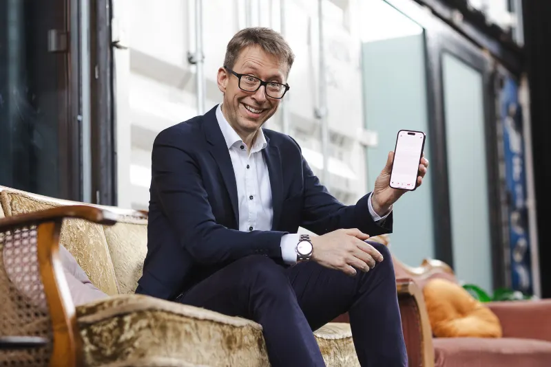 Daniel Schwenger im Anzug sitzt auf einem Sofa und hält lächelnd ein Smartphone